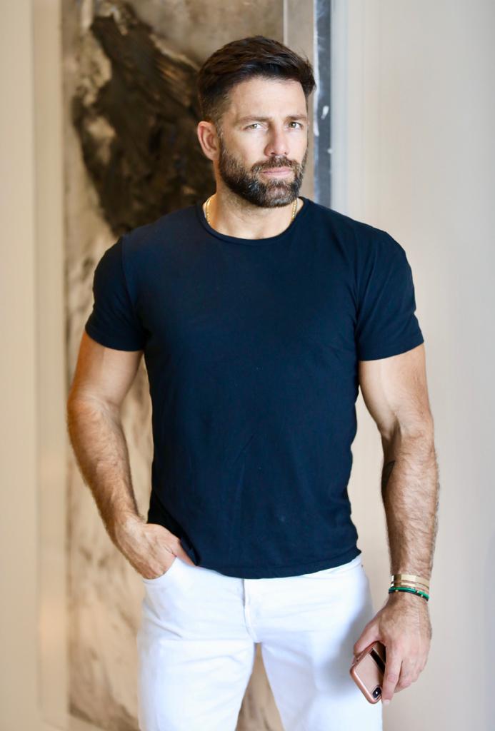 Man in a black t-shirt and white pants standing indoors.