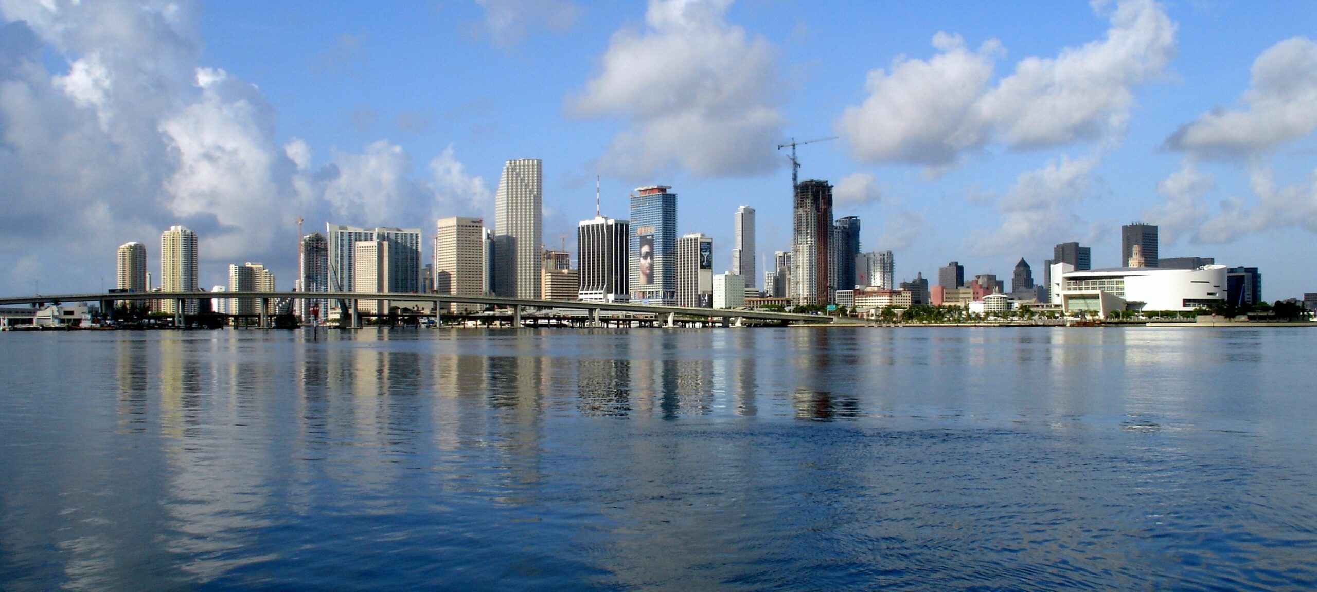City skyline with tall buildings reflecting on calm water under a blue sky.