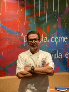 Man in white chef coat standing with arms crossed in front of colorful graffiti wall.