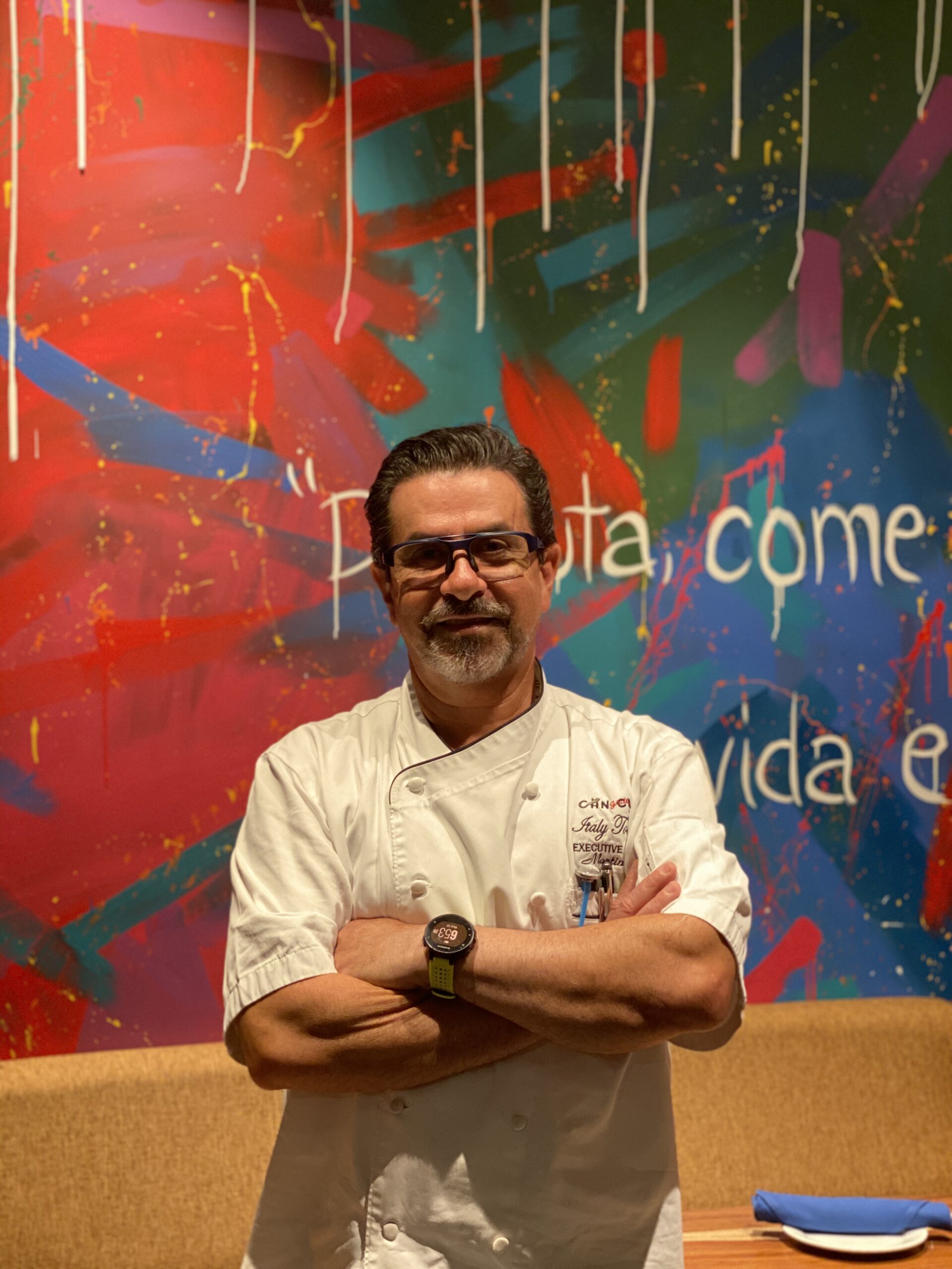 Man in white chef coat standing with arms crossed in front of colorful graffiti wall.