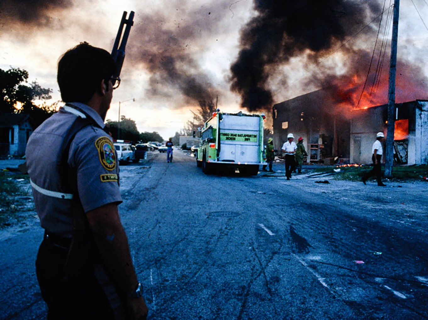 A person watches a street engulfed in flames and smoke, with emergency vehicles nearby.