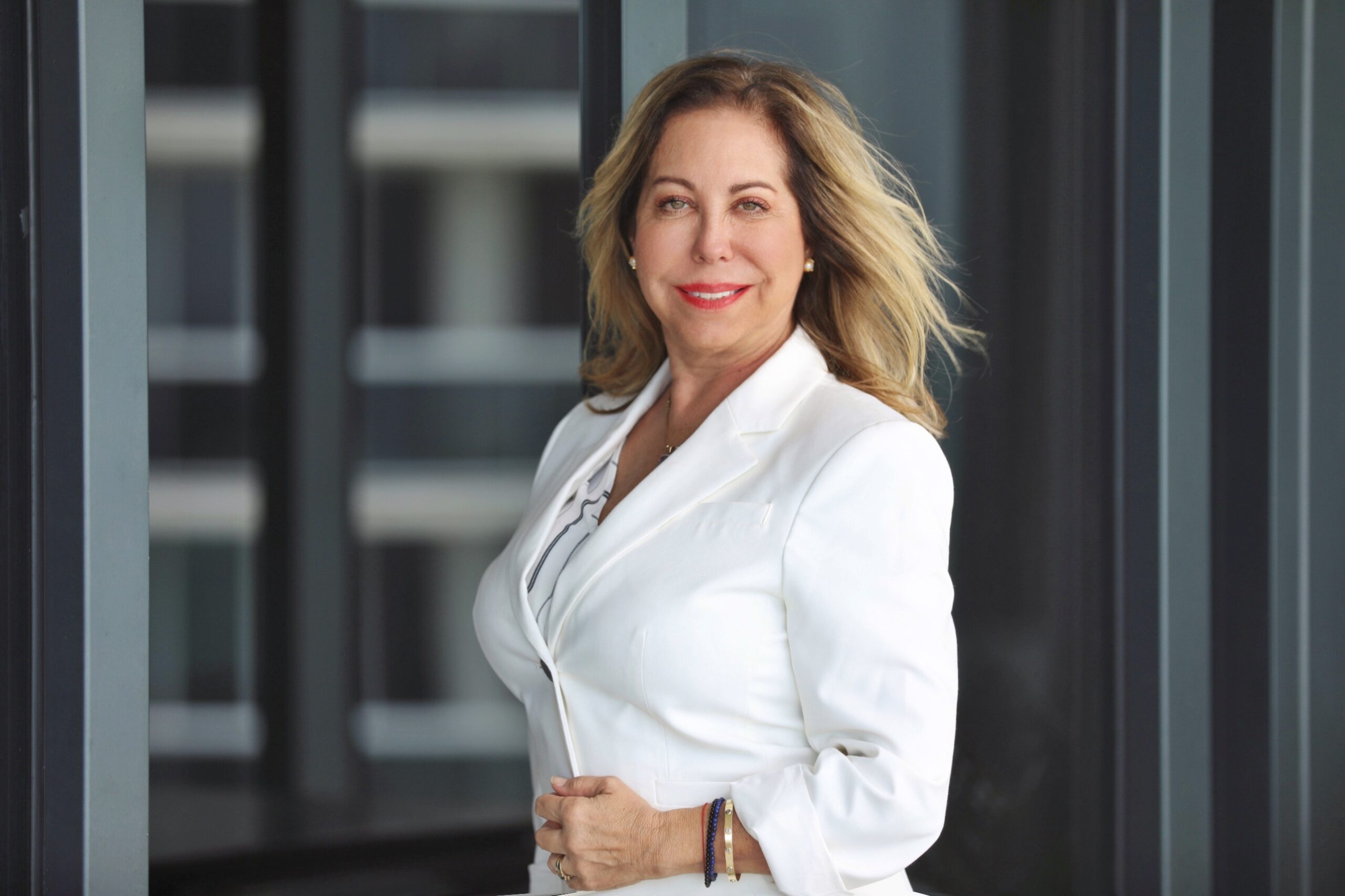 Confident woman in a white blazer poses with a smile against a modern office background.