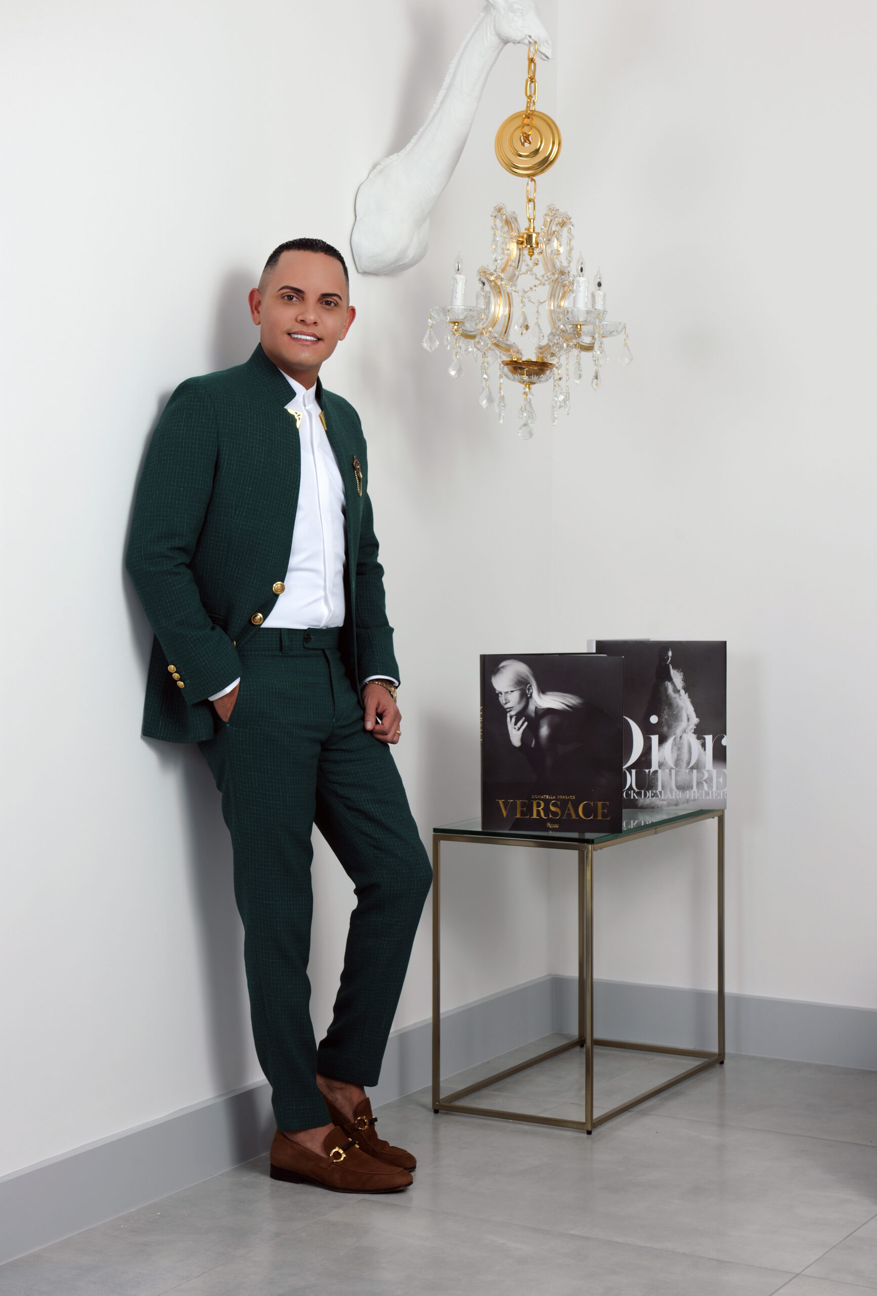 A man in a green suit smiling beside a chandelier and black-and-white artwork.