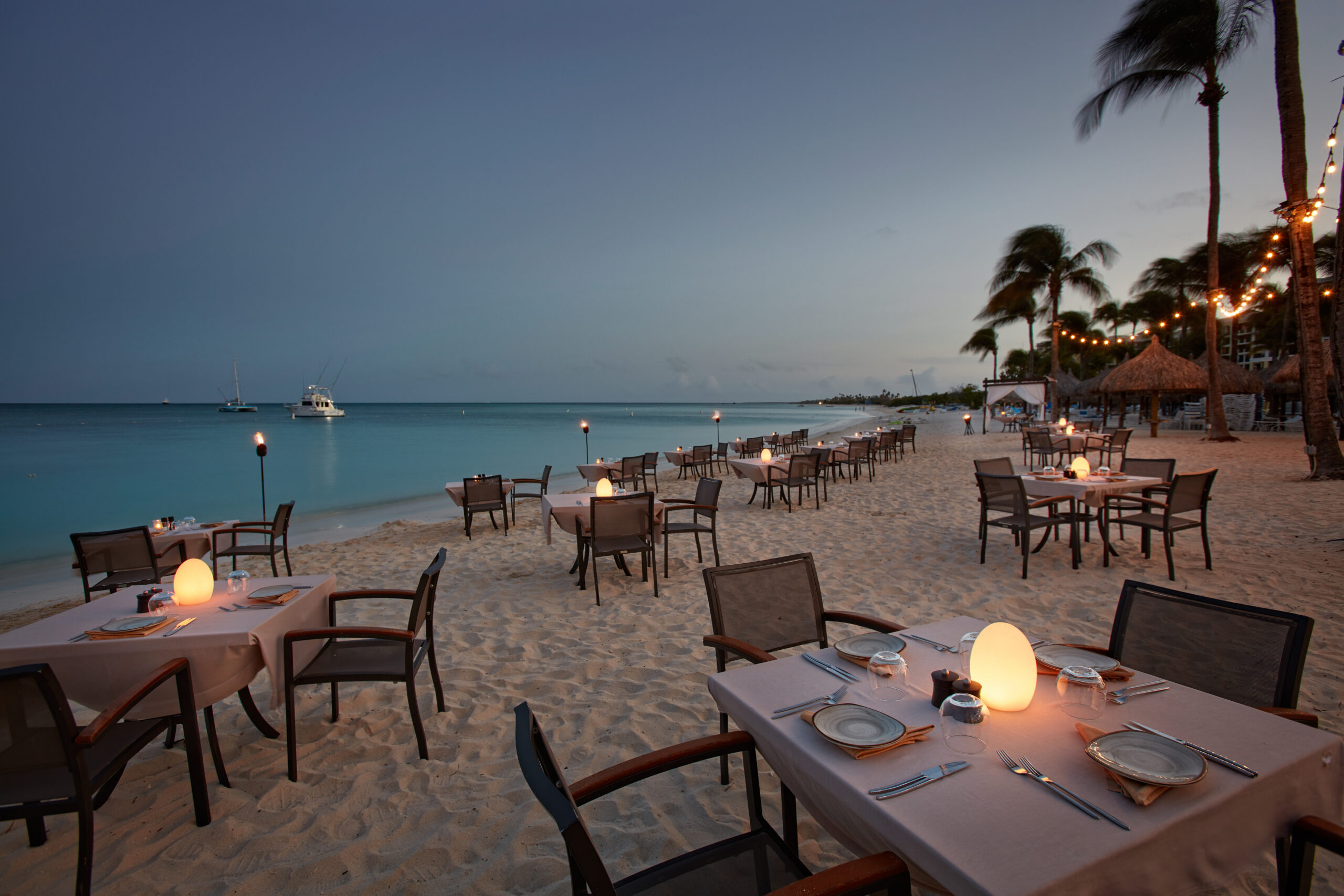 Elegant beachside dining setup at dusk with tables and chairs on sand.