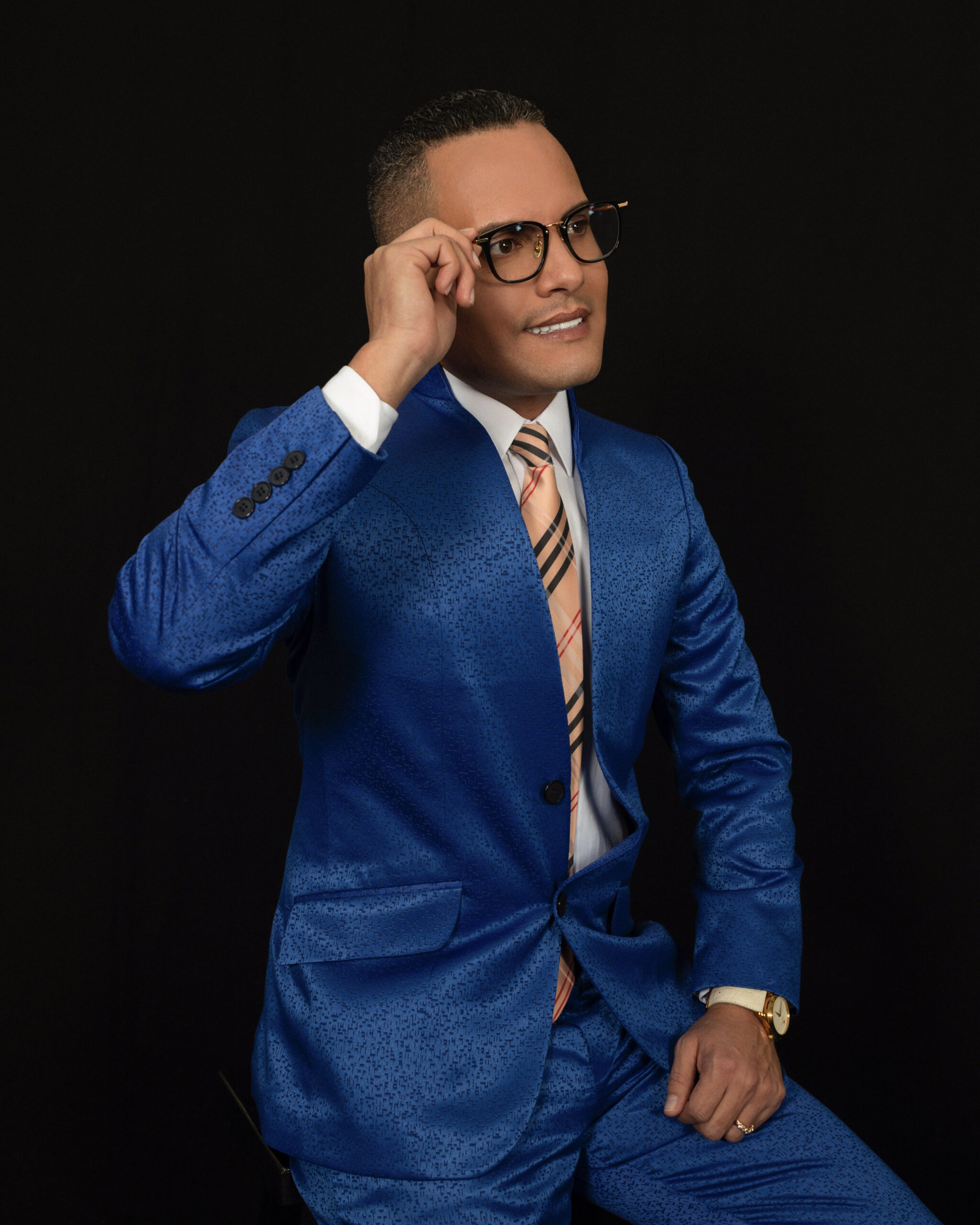 Man in blue suit adjusting glasses with a confident smile.