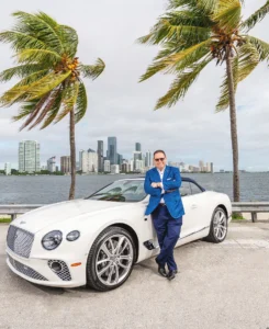 Man in blue suit leaning on a white luxury car by the waterfront with cityscape background.