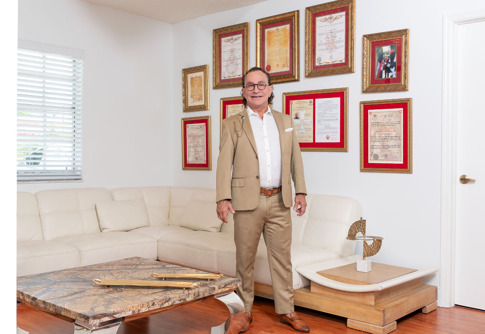 A man in a tan suit stands in a stylish living room with framed certificates on the wall.