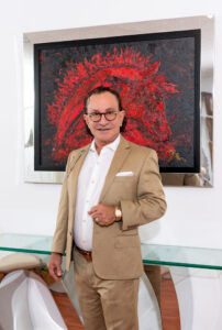 Distinguished man in a beige suit stands confidently indoors against a red artwork backdrop.