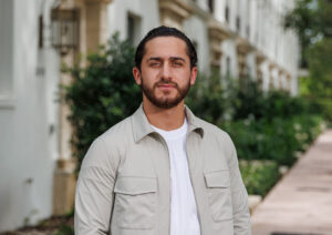 Man with beard standing outdoors in casual light jacket and white shirt.