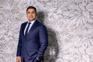 Confident man in a navy suit poses against a textured wall.