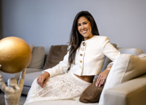 A confident woman in a stylish white outfit sitting on a sofa, smiling warmly.