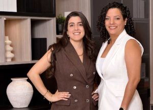 Two confident women posing indoors, dressed in professional attire.