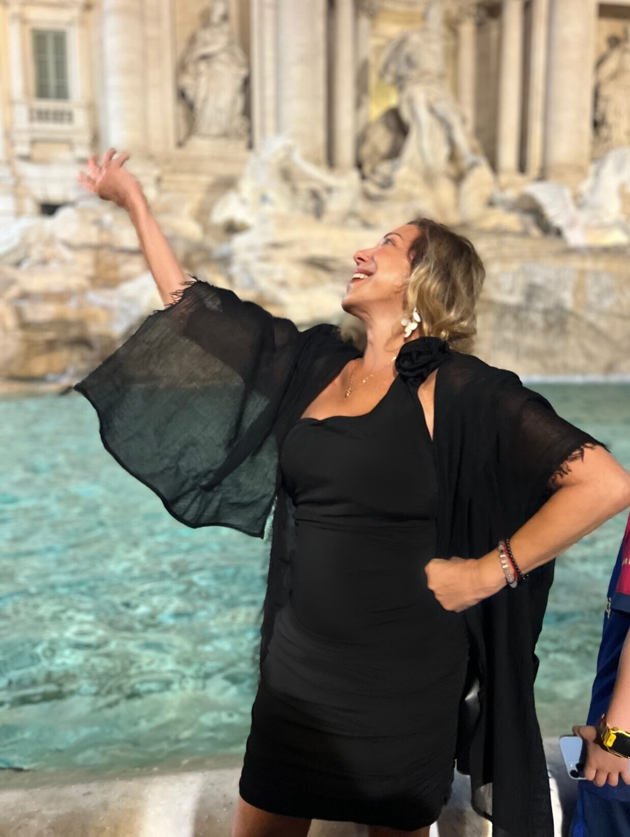 Woman in black dress joyfully posing by a fountain.