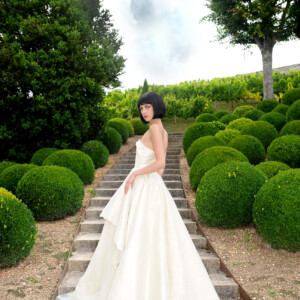 Bride in elegant white gown stands on garden steps surrounded by manicured bushes.
