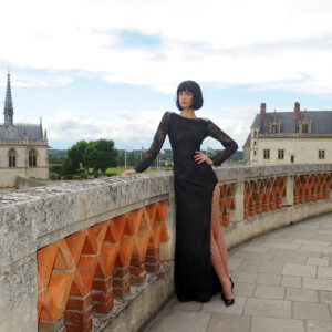 Woman in elegant black dress poses on historic stone balcony with scenic backdrop.