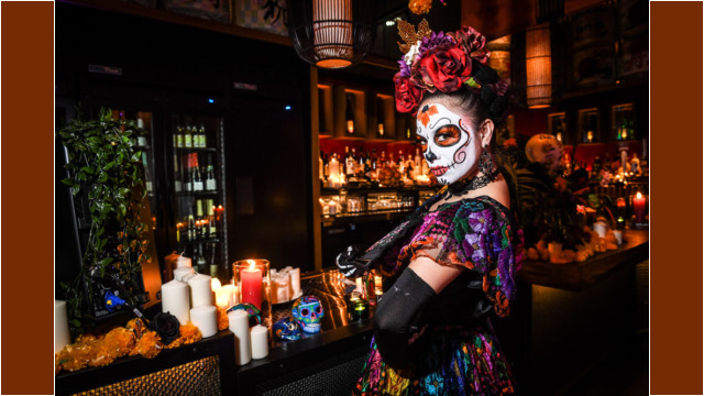 Woman in vibrant Day of the Dead makeup and attire at a candlelit bar.