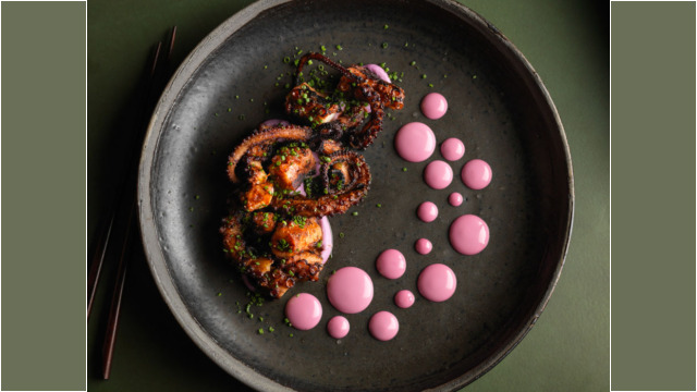 Grilled octopus with pink sauce dots on a black plate.