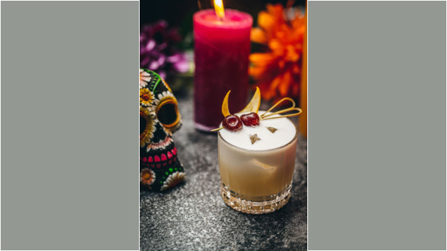 A creamy cocktail garnished with cherries beside a colorful sugar skull decoration.