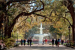 People stroll near a large fountain under autumn trees in a park.