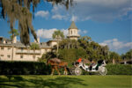 Horse-drawn carriage rides near a grand estate under a blue sky.