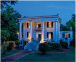 Elegant historic mansion illuminated at dusk with grand columns and lush greenery.