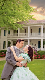 Couple embracing in front of a large white house with a porch.