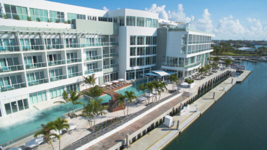 Modern waterfront hotel with pool and palm trees along a marina.