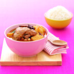 A bowl of chicken stew with rice in the background.