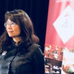 A woman with glasses stands thoughtfully against a red and white backdrop.