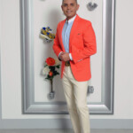 A man in a bright orange blazer posing indoors against a decorative wall.