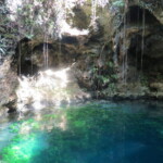 A clear blue cenote surrounded by rocky walls and hanging vines.