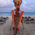 A colorful skeleton figure stands on a beach with empty lounge chairs and ocean in the background.