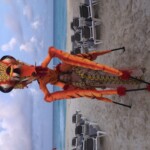 A colorful skeleton figure stands on a beach with empty lounge chairs and ocean in the background.