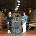 Two women posing beside a large Buddha statue indoors.