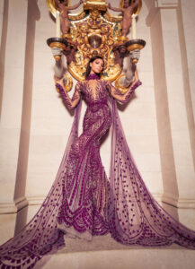 Elegant woman in a detailed purple gown posing gracefully indoors.