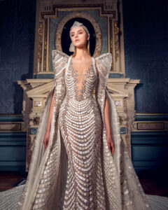 A woman in an elegant, intricate gown with a dramatic cape poses in a regal setting.