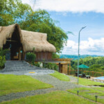 Rustic thatched-roof cottage with stone steps amid lush greenery under a blue sky.