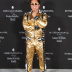 Man in a shiny gold tracksuit posing with crossed arms and sunglasses.