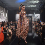Model walking down runway in a flowing, patterned dress at a fashion show.