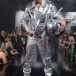 Model in a shiny silver space-themed outfit holding a matching helmet on runway.