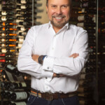 Smiling man in white shirt stands confidently in front of wine racks.