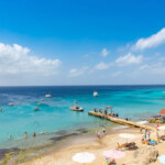 Sunny beach with clear blue water and boats near a wooden pier.