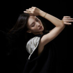 Woman gracefully posing with arms raised against a dark background.