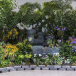 A serene Buddha statue surrounded by vibrant flowers and lush greenery.