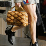 Person holding a quilted tan handbag and wearing black ankle boots.
