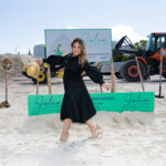 A woman in a black dress poses on sandy ground at a construction site with machinery in the background.