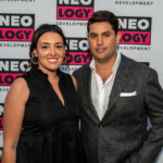 A woman and a man posing at a Neology event with branded backdrop.