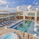 Luxurious cruise ship pool area with lounge chairs and hot tubs under a blue sky.