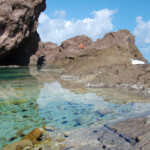 Clear turquoise water laps against rugged coastal rocks under a bright blue sky.