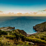 Scenic coastal view with grassy hills and calm ocean under a colorful sky.